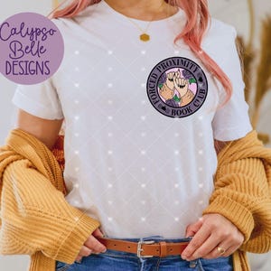 May include: White t-shirt with a round graphic that says "FORCED PROXIMITY BOOK CLUB" with two hands and roses. The shirt has a diamond pattern and the brand "Calypso Belle Designs" is on the left side. A mustard-colored sweater is draped over the shoulders.