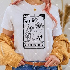 May include: A white t-shirt with a black and white illustration of a tarot card. The card is labeled "The Bride" and features a hand with a ring on the middle finger, surrounded by roses.