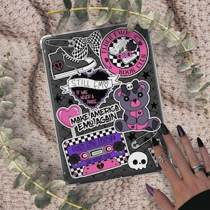 May include: A clear tablet case decorated with various stickers. The stickers include a black and white checkered bow, a gray high-top sneaker, a heart-shaped balloon, and a teddy bear. Text includes "STILL EMO" and "MAKE AMERICA EMO AGAIN".