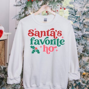 May include: White sweatshirt with a green holly leaf and red berry design. The text "Santa's favorite ho" is printed in red, green, and pink.