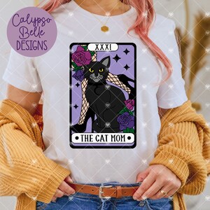 May include: A white t-shirt featuring a purple tarot card design with a black cat sitting on a woman's leg. The card reads "XXXI The Cat Mom".
