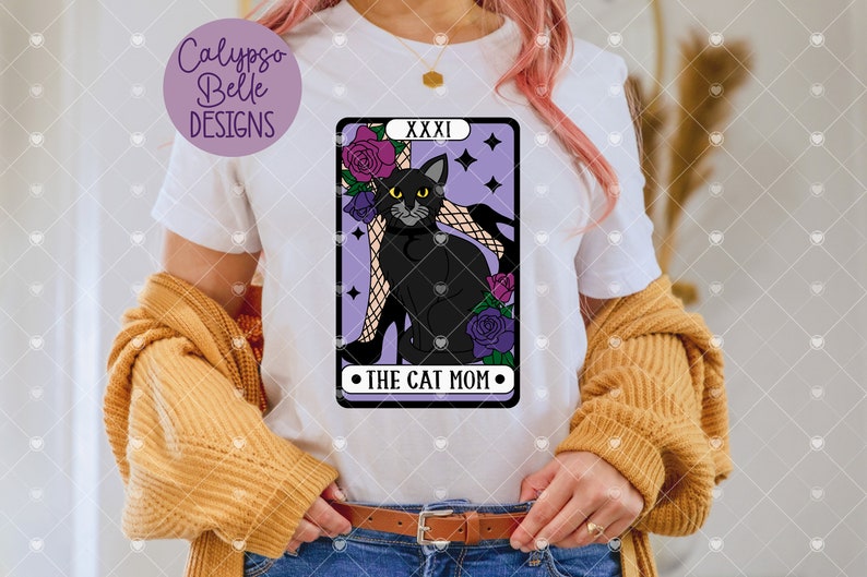 May include: A white t-shirt featuring a purple tarot card design with a black cat sitting on a woman's leg. The card reads "XXXI The Cat Mom".