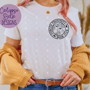 May include: White t-shirt featuring a black and white graphic design of a circle with the text "Forced Proximity Book Club" and a design of two hands holding a book.