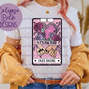Puede incluir: Camiseta blanca con un dise&ntilde;o de carta de tarot rosa y morado. La carta presenta un coraz&oacute;n con una cuerda alrededor y dos manos que se sostienen. El texto en la carta dice "Let's Pretend" y "Fake Dating".