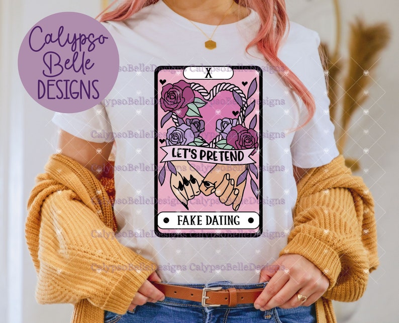 Puede incluir: Camiseta blanca con un dise&ntilde;o de carta de tarot rosa y morado. La carta presenta un coraz&oacute;n con una cuerda alrededor y dos manos que se sostienen. El texto en la carta dice "Let's Pretend" y "Fake Dating".