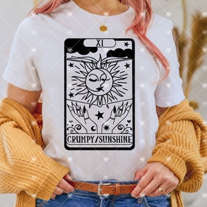 May include: A white t-shirt with a black and white illustration of a tarot card. The card is the "XI" card, and it features a sun and moon with a face, and the text "GRUMPY/SUNSHINE".