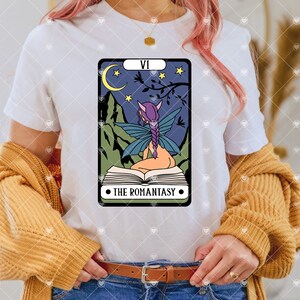 May include: White t-shirt with a graphic of a tarot card featuring a fairy reading a book. The card is labeled "VI THE ROMANTASY".