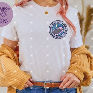 May include: White t-shirt featuring a circular graphic with the text "Only One Bed Book Club" and a bed and moon illustration. The shirt is paired with a mustard-coloured sweater and blue jeans. The brand name "Calypso Belle Designs" is in a purple circle.