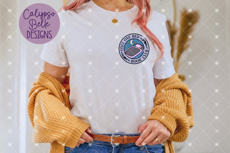 May include: White t-shirt featuring a circular graphic with the text "Only One Bed Book Club" and a bed and moon illustration. The shirt is paired with a mustard-coloured sweater and blue jeans. The brand name "Calypso Belle Designs" is in a purple circle.