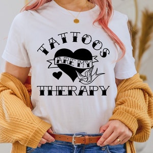 Op de afbeelding: Wit T-shirt met een zwarte afbeelding met de tekst "Tattoos zijn mijn therapie" met een hart en een vogelontwerp.
