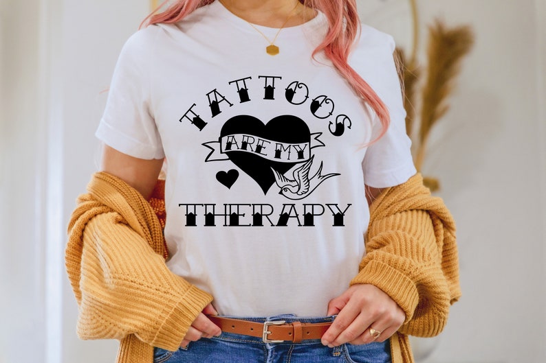 Op de afbeelding: Wit T-shirt met een zwarte afbeelding met de tekst "Tattoos zijn mijn therapie" met een hart en een vogelontwerp.
