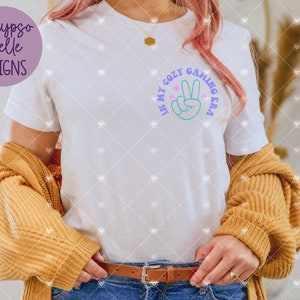 May include: White t-shirt with a graphic of a peace sign and the text "In my cozy gaming era".