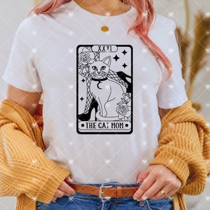 Puede incluir: Camiseta blanca con una ilustraci&oacute;n en blanco y negro de un gato con un collar y sentado en un zapato de tac&oacute;n alto. La ilustraci&oacute;n est&aacute; en el estilo de una carta de tarot y dice "XXXI The Cat Mom".