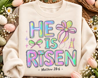 May include: A cream-colored crewneck sweatshirt featuring the text HE IS RISEN and Matthew 28:6 in a holographic, bubble-letter style. The design includes a large pink bow, a small green bow, and a yellow bunny silhouette graphic.
