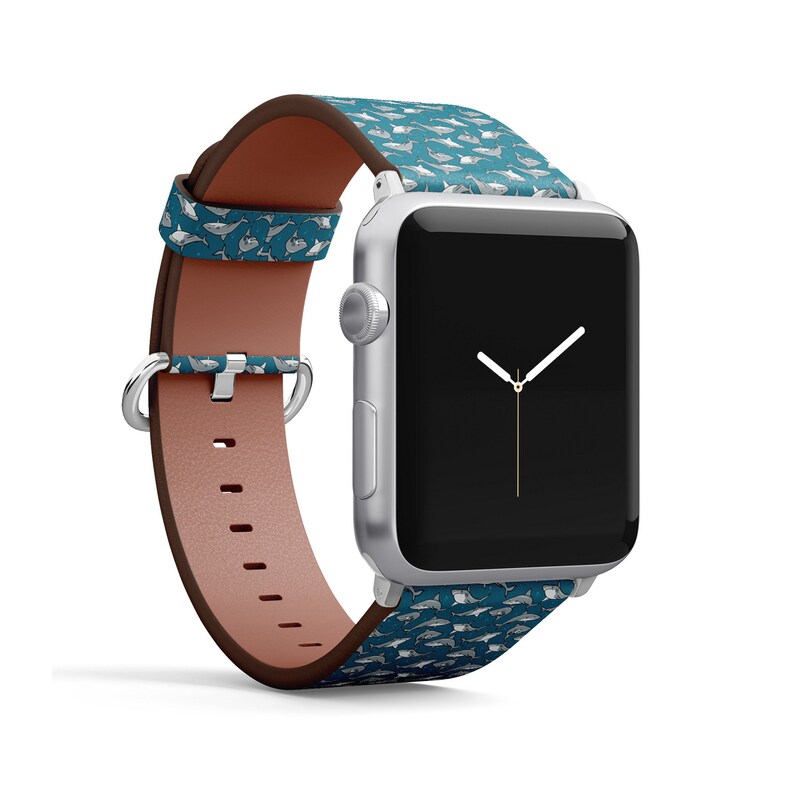 May include: A silver Apple Watch with a teal and white leather band featuring a repeating pattern of sharks. The watch face is black with white hands.
