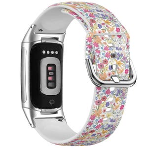 Puede incluir: Una pulsera de reloj de silicona blanca con un patrón floral. La pulsera tiene una hebilla plateada y es compatible con un rastreador de actividad física.