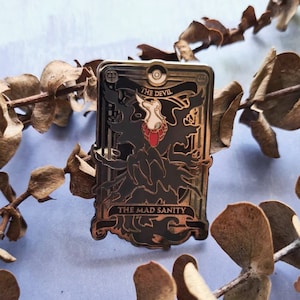 Könnte beinhalten: Goldene und schwarze Emaille-Anstecknadel mit einem Tarotkarten-Design mit dem Text "The Devil" und "The Mad Sanity".