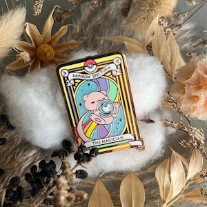 Puede incluir: Pin rectangular con un personaje de dibujos animados sosteniendo una bola de cristal, con el texto "Rainbow Specturm" y "The Magician". El pin tiene un borde color cobre y está colocado sobre un fondo de flores secas y algodón.