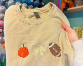 Könnte beinhalten: Ein beiger Pullover mit gestickten Bildern von einem Kürbis, einer Schleife und einem Fußball. Der Text "WHITE PINE craft co." ist unten auf dem Pullover gestickt.