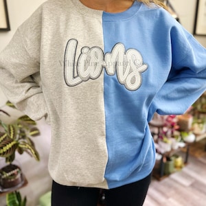 May include: A two-toned sweatshirt, split vertically with gray on the left and light blue on the right. The word "Lions" is embroidered across the chest in a white and silver script. The sweatshirt is a crew neck style.