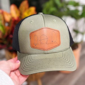 May include: Olive green and black trucker hat with a brown leather patch that reads "DAD". The hat has a curved brim and a mesh back. The text "The Pine Apparel Co." is below the word "DAD".