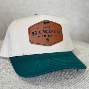 May include: A white and green baseball cap with a brown leather patch that says "Talk Birdie To Me" with a golf ball icon.