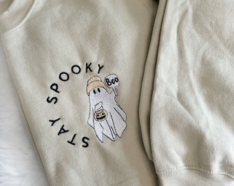 Könnte beinhalten: Ein hellbeiger Pullover mit einem gestickten Design eines Geistes, der einen Kürbis-Eimer hält. Der Geist trägt einen Hut und hat eine Sprechblase, in der "Boo" steht. Der Text "STAY SPOOKY" ist in einem Kreis um den Geist geschrieben.