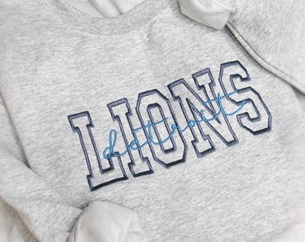 Può includere: Una felpa grigio chiaro con la scritta "LIONS" ricamata in lettere maiuscole blu navy con un "Detroit" in corsivo blu. La felpa ha un girocollo e maniche lunghe. Il logo White Pine Craft Co. è visibile.