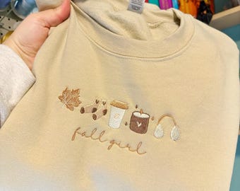 May include: A beige sweatshirt with embroidered fall-themed imagery, including a leaf, socks, a coffee cup, a candle, and earmuffs. The text "fall girl" is embroidered below the imagery. The sweatshirt has a white tag with the text "White Pine craft co."