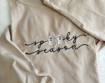 Könnte beinhalten: Ein beigefarbenes Langarmshirt mit gerippter Struktur. Das Shirt hat ein gesticktes Design mit zwei Geistern und dem Text "Spooky Season".