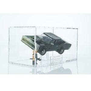 Könnte beinhalten: Ein schwarzes Dodge Charger Modellfahrzeug in einer transparenten Acryl-Vitrine. Das Auto ist schräg gestellt und der Text "Fast & Furious Dodge Charger '70" ist auf dem Sockel der Vitrine sichtbar.