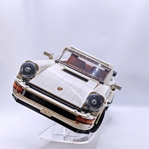 May include: A white Lego Porsche 911 Turbo model car on a clear acrylic display stand. The car has black headlights and a red Porsche logo on the front.