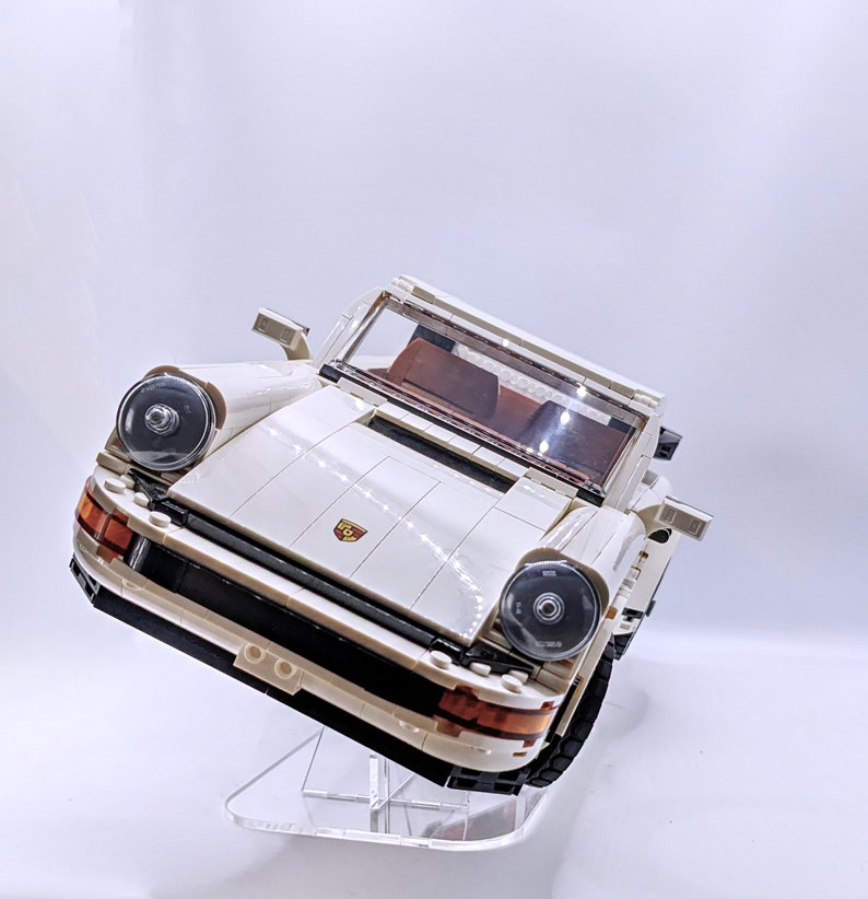 May include: A white Lego Porsche 911 Turbo model car on a clear acrylic display stand. The car has black headlights and a red Porsche logo on the front.