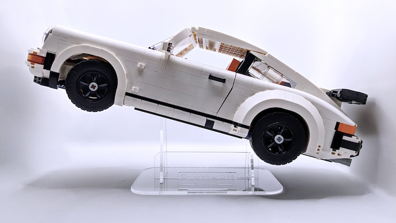 May include: A white Lego Porsche 911 Turbo model car on a clear acrylic display stand. The car is tilted at an angle, with the front wheels off the ground.