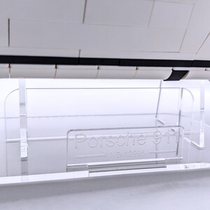May include: A clear acrylic display stand for a white Lego Porsche 911 model car. The stand has a rectangular base and two vertical supports. The base has the text "Porsche 911" printed on it.