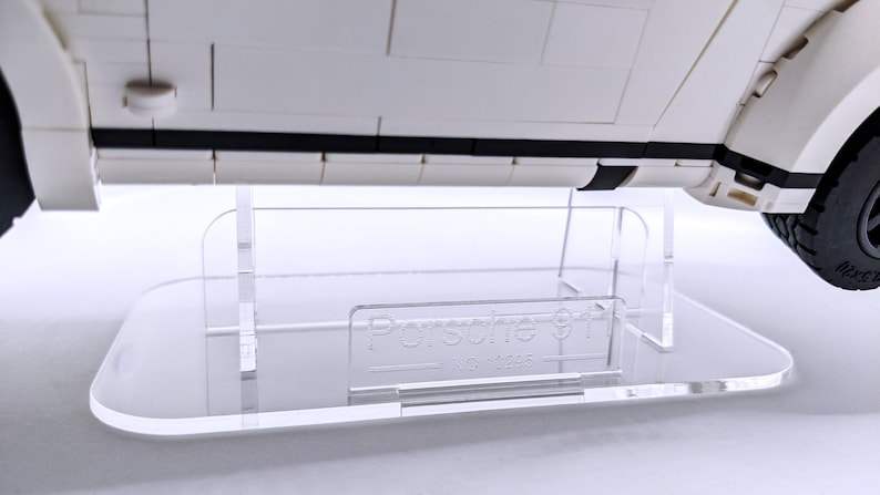 May include: A clear acrylic display stand for a white Lego Porsche 911 model car. The stand has a rectangular base and two vertical supports. The base has the text "Porsche 911" printed on it.