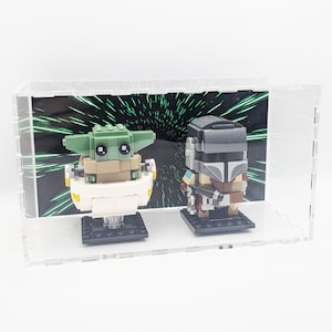 Könnte beinhalten: Eine transparente Acryl-Vitrine mit grünem und schwarzem Hintergrund, die eine Lego-Minifigur von The Mandalorian und eine Lego-Minifigur von Grogu, auch bekannt als Baby Yoda, in einer schwebenden Kapsel zeigt.