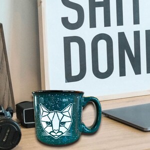 May include: A framed poster with the text "GET SHIT DONE."  A teal blue mug with a white geometric cat design sits on a wooden table in front of the poster.