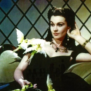 May include: A woman with dark hair and a black dress sits on a couch with a group of small, light green birds perched on her shoulder. She is wearing a gold necklace with a large, dark gemstone pendant and a gold bracelet on her right wrist.