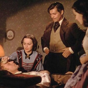 May include: A scene from a movie with three people in a room. A man in a suit stands next to a woman in a blue and white striped dress. The woman is tending to a man lying in bed. The room is dimly lit with a lamp on a table in the foreground.