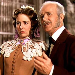 May include: A woman in a brown dress with a white lace collar and a purple ribbon stands next to a man in a dark suit and white shirt. The woman is looking to the side, while the man is looking at the camera and smiling.