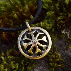 May include: A brass pendant necklace with a circular design featuring a stylized eight-pointed star. The pendant is on a black leather cord and is resting on a bed of green moss.