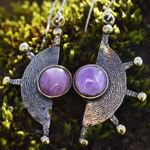 Könnte beinhalten: Ein Paar Ohrringe mit einem bronzefarbenen Halbmond-Design und einem violetten Edelstein in der Mitte. Die Ohrringe haben eine strukturierte Oberfläche und goldene Akzente.
