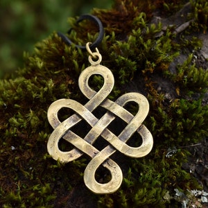 May include: A gold-colored metal pendant in the shape of a Celtic knot. The pendant is hanging from a black cord and is resting on a bed of green moss.