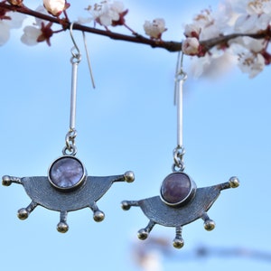 Könnte beinhalten: Ein Paar silberne Ohrringe mit geometrischem Design und einem violetten Stein in der Mitte. Die Ohrringe hängen an einem Zweig mit weißen Blüten.