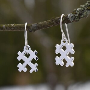 Op de afbeelding: Een paar zilveren oorbellen met een kruisvormig ontwerp. De oorbellen hangen aan haken en worden getoond tegen een vage natuurlijke achtergrond. De oorbellen zijn gemaakt van glanzend, reflecterend metaal.