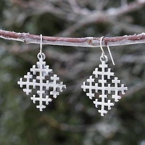 Op de afbeelding: Een paar zilveren oorbellen met een geometrisch kruisontwerp. De oorbellen hangen aan een tak.