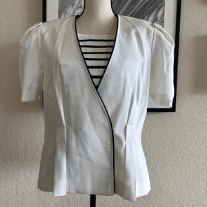 May include: A white linen blouse with a black trim and a striped neckline. The blouse has a wrap-style front and a fitted silhouette.