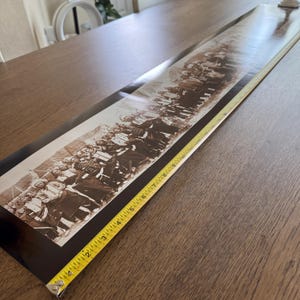 May include: A long, rectangular sepia-toned panoramic photograph featuring a large group of people in historical attire, positioned next to a yellow measuring tape that indicates a length of approximately 24 inches along the bottom edge.
