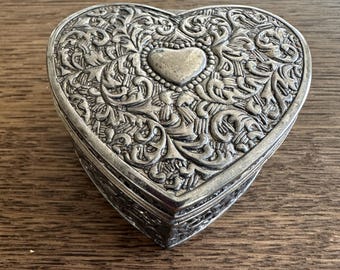 Puede incluir: Una caja de corazón de plata con un diseño ornamentado. La caja tiene un corazón en relieve en el centro y está decorada con intrincados patrones florales.